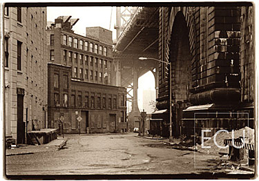 Black and white photo rendered in sepia of the Manhattan Bridge shot from the Brooklyn side of the East River showing piers and support arches